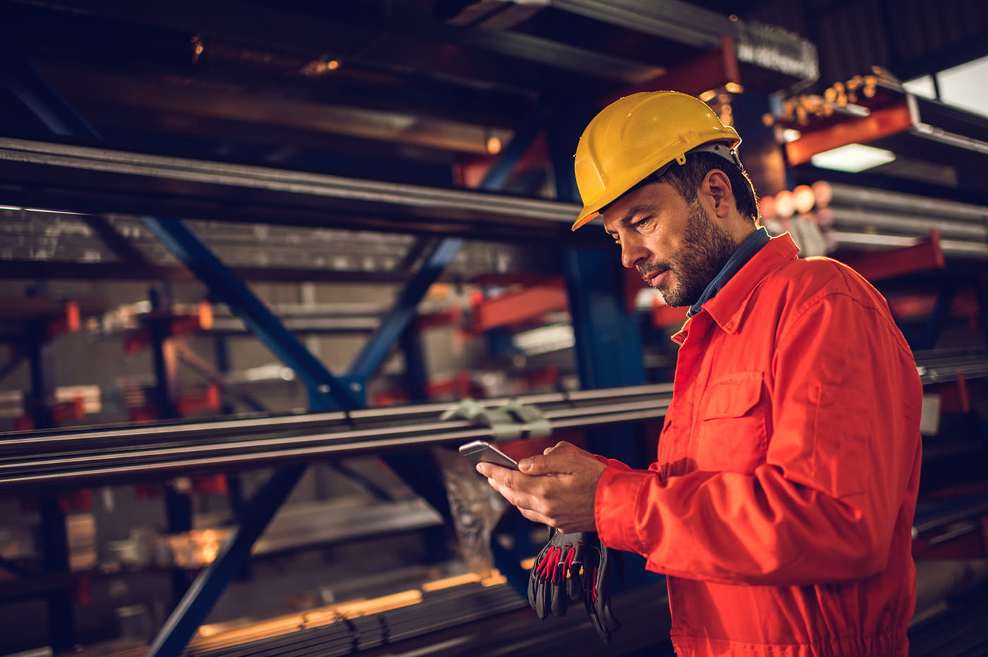 A man in a hardhat and jumpsuit looking at his cellphone near some pipes.