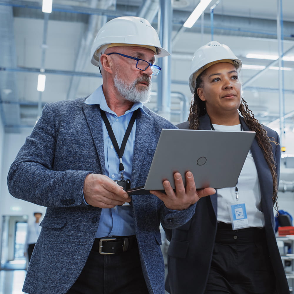 A man holding a laptop and talking to woman, both wearing construction helmets.